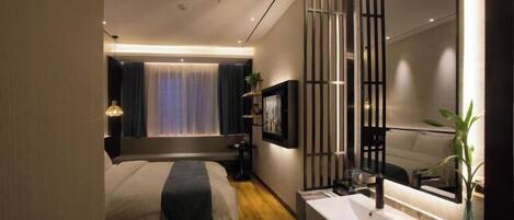 A modern hotel room with a large bed, a bedside table, a mirror, a bathroom with a sink, and a window with curtains.