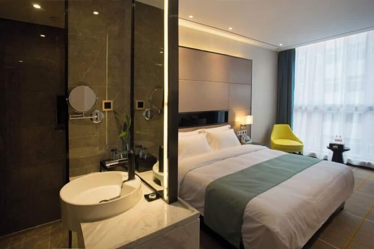 Una habitación de hotel moderna con una cama grande, un baño con lavabo y espejo, una mesita de noche y una ventana con cortinas.