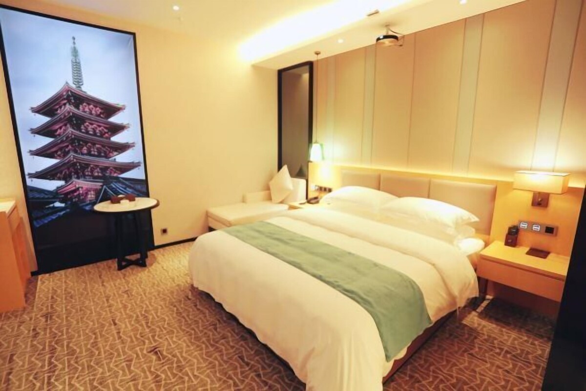 A hotel room with a bed, bedside tables, a nightstand, a wall-mounted lamp, and a large poster of a pagoda.