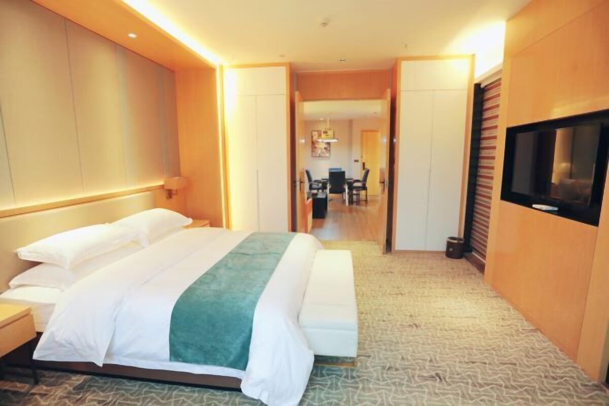 A hotel room with a bed, bedside table, TV, and a view into the dining area.