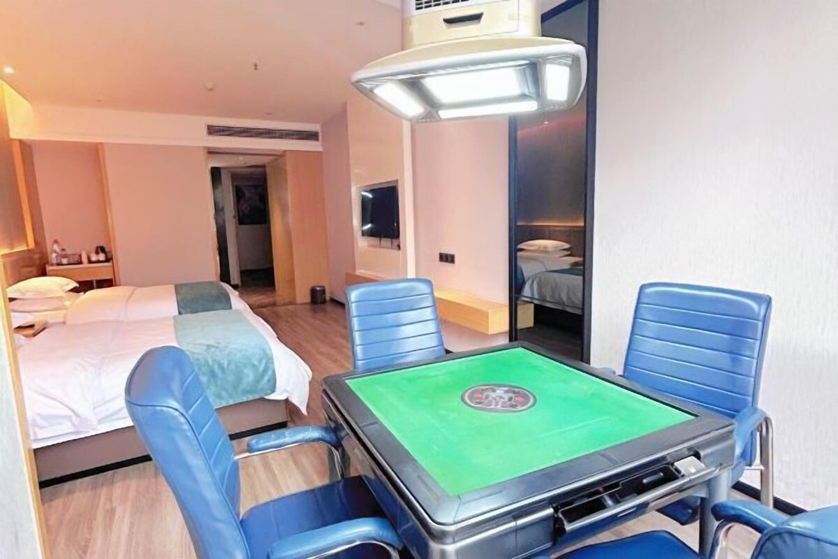 A hotel room with a bed, a desk with a green table, and blue chairs.