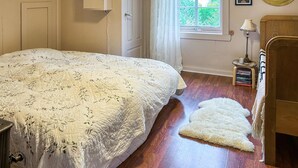 Une chambre avec un grand lit, un plancher en bois, une petite table de chevet avec des livres, une lampe et une fenêtre donnant sur un paysage verdoyant.