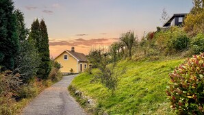 Une route de campagne mène à une maison jaune, dont la porte est ornée du numéro 30, et qui est entourée de verdure et d’arbres.