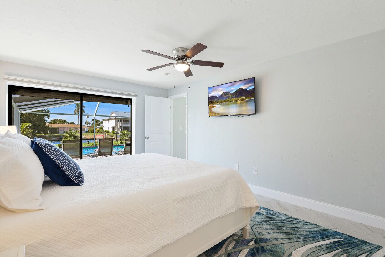A bedroom with a bed, a ceiling fan, a painting, and a view of a pool and buildings.