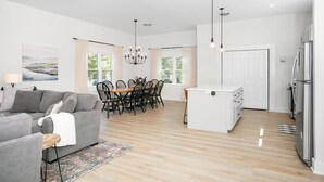 A modern kitchen with white cabinets and a built-in island, a dining area with a wooden table and chairs, and a living room with a grey sofa and a rug.