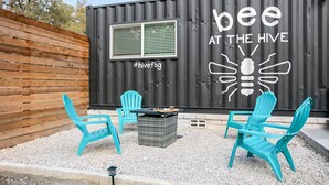 A black container with "bee at the hive" and a bee graphic, outdoor seating with turquoise chairs, and a small table.