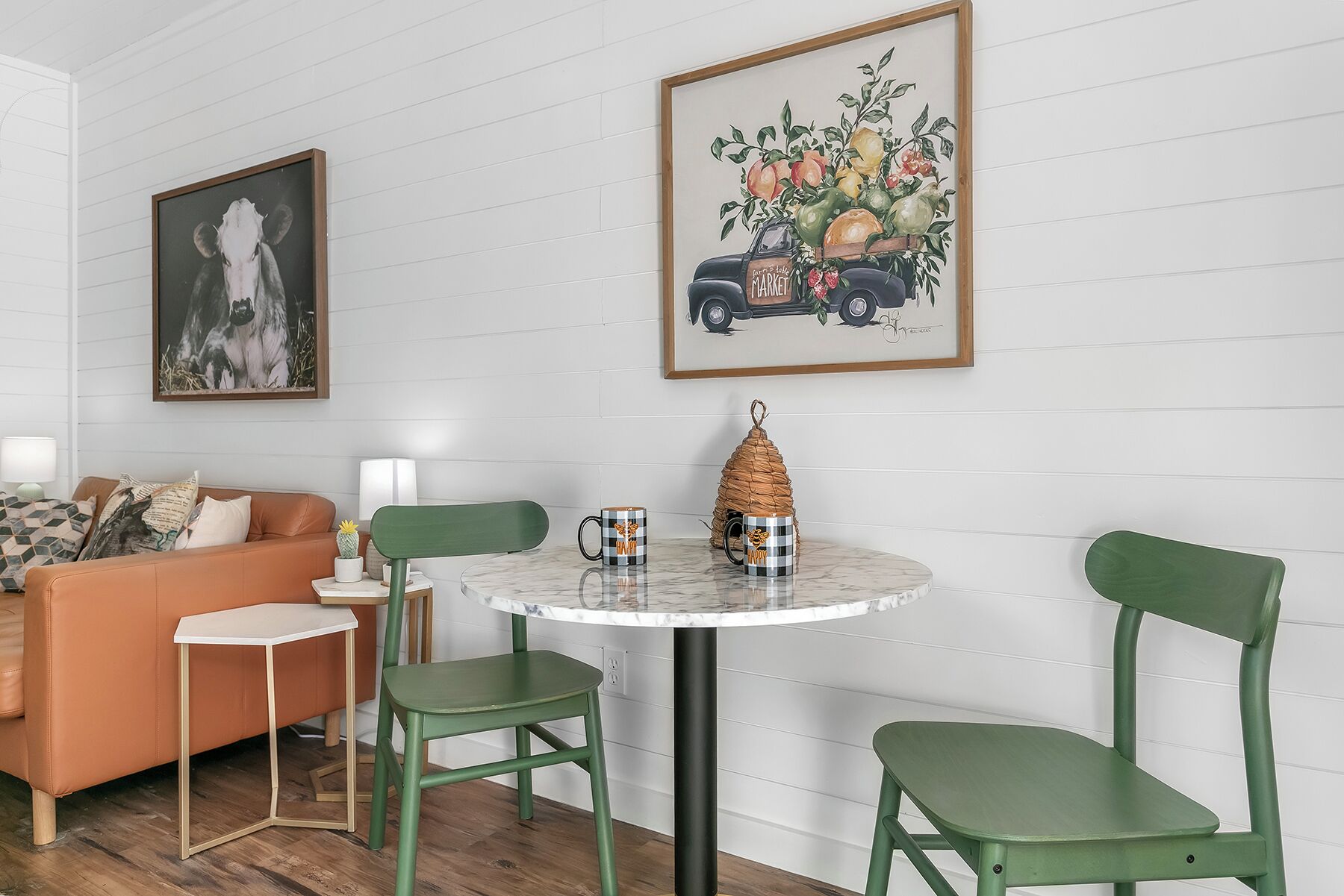 Un salón acogedor con un sofá verde, una pequeña mesa redonda y dos sillas verdes. Hay dos obras de arte enmarcadas en la pared, una que representa una vaca y otra un coche antiguo con fruta.