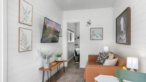 A modern living room with a sofa, a coffee table, and a TV. There are framed pictures on the walls and a plant on a side table. - Honey at the Hive, 1/1 | Private Hot Tub | Gas Fire Pit | Cornhole (Fredericksburg)