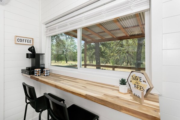 A kitchen with a coffee station, a sign reading "SAVE the BEES," and a view of trees outside.