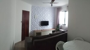 Ein Wohnzimmer mit einer Couch, einem an einer strukturierten Wand montierten Fernseher, einem Esstisch mit Stühlen und einem Deckenventilator.