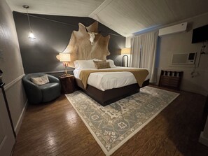 A bedroom with a bed, a nightstand, a lamp, a wall-mounted TV, and an air conditioning unit. - The Ranch House Motel – Your West Texas Getaway Near San Angelo -  Mesquite Room (Christoval)
