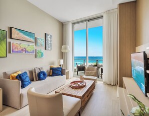 A modern living room with a sofa, coffee table, and artwork on the wall. A large window offers a view of the ocean.