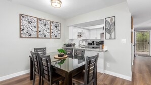 A dining area with a glass table, dark chairs, and a potted plant. There are white cabinets and a kitchen area in the background. -  NEW & GORGEOUS Lakefront ZEN 3 bedrooms [2 KING +1 Queen] (Phoenix)