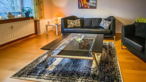A living room with a glass coffee table, a sofa, and a chair. There is a painting on the wall and a window with curtains.