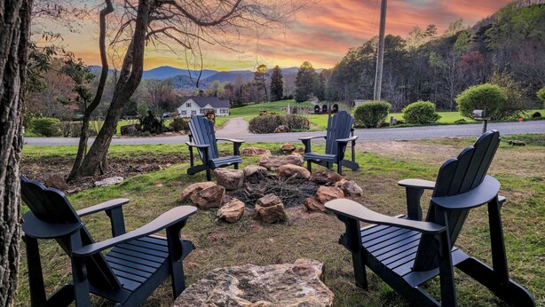A serene outdoor seating area with Adirondack chairs and a fire pit, overlooking a scenic landscape with a house and mountains in the distance.