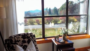 A room with a large window offering a view of a garden and trees. There is a cow-print chair and a small table with a plant and a sign that reads "Farm Fresh."