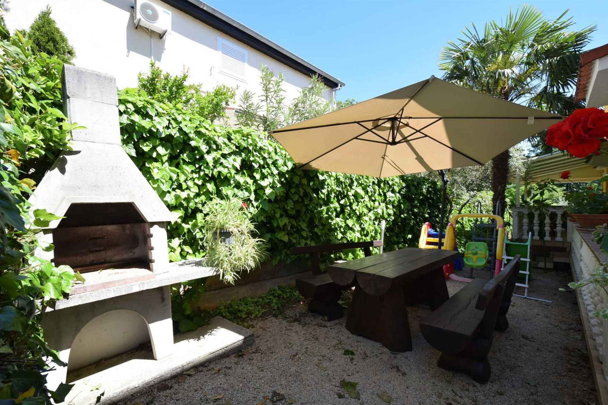 En udendørs terrasse med en støbejernsgrill, et træbord og en stor parasol, der giver skygge.