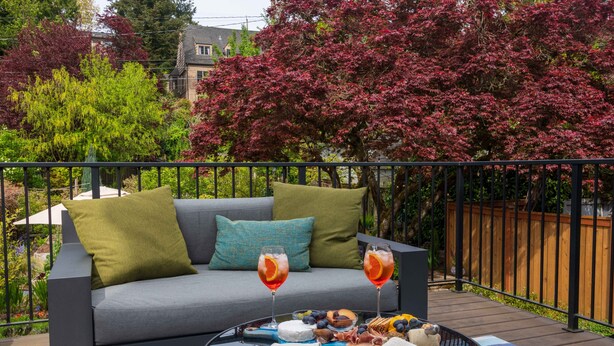 A balcony with a grey sofa, green and blue cushions, a round table with a glass top, and a variety of food and drinks.