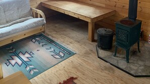 A wooden cabin interior with a sofa, a bench, a fireplace, and a window overlooking a forest.