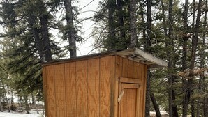 A wooden shed with a door and a small overhang, surrounded by trees and snow on the ground.