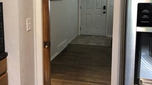 Un pasillo con piso de madera, una puerta blanca al final y un refrigerador a la derecha.