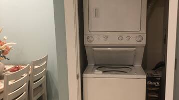 A compact laundry room with a front-loading washer and dryer, a dining table with chairs, and a partially visible door leading to another room.