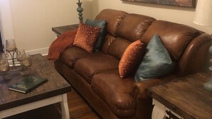 Un salón acogedor con un sofá de cuero marrón, una mesa de centro de madera y un cuadro de un paisaje forestal.