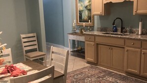 Un comedor con una mesa, sillas y un tapete. Se observa una cocina con armarios y un lavamanos.