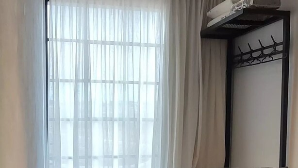 A bedroom with a bed, a small stool, a window with curtains, and a wall-mounted shelf.