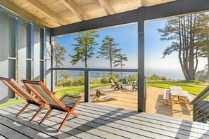 A covered outdoor deck with wooden flooring, lounge chairs, and a view of a grassy area with trees and a body of water.
