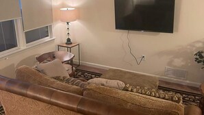Ein Wohnzimmer mit braunem Sofa, einem an der Wand montierten Fernseher, einer Lampe auf einem Beistelltisch und einem Fenster mit Jalousien.