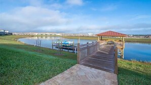Un muelle de madera que conduce a un pabellón junto a un lago tranquilo, con barcos amarrados cerca y un puente al fondo.