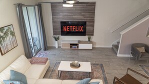 Una sala de estar con un sofá blanco, una mesa de centro, un televisor montado en una pared de madera y una escalera al fondo.