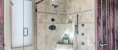 A walk-in shower with a glass door, stone tiles, and a wooden shelf with toiletries.
