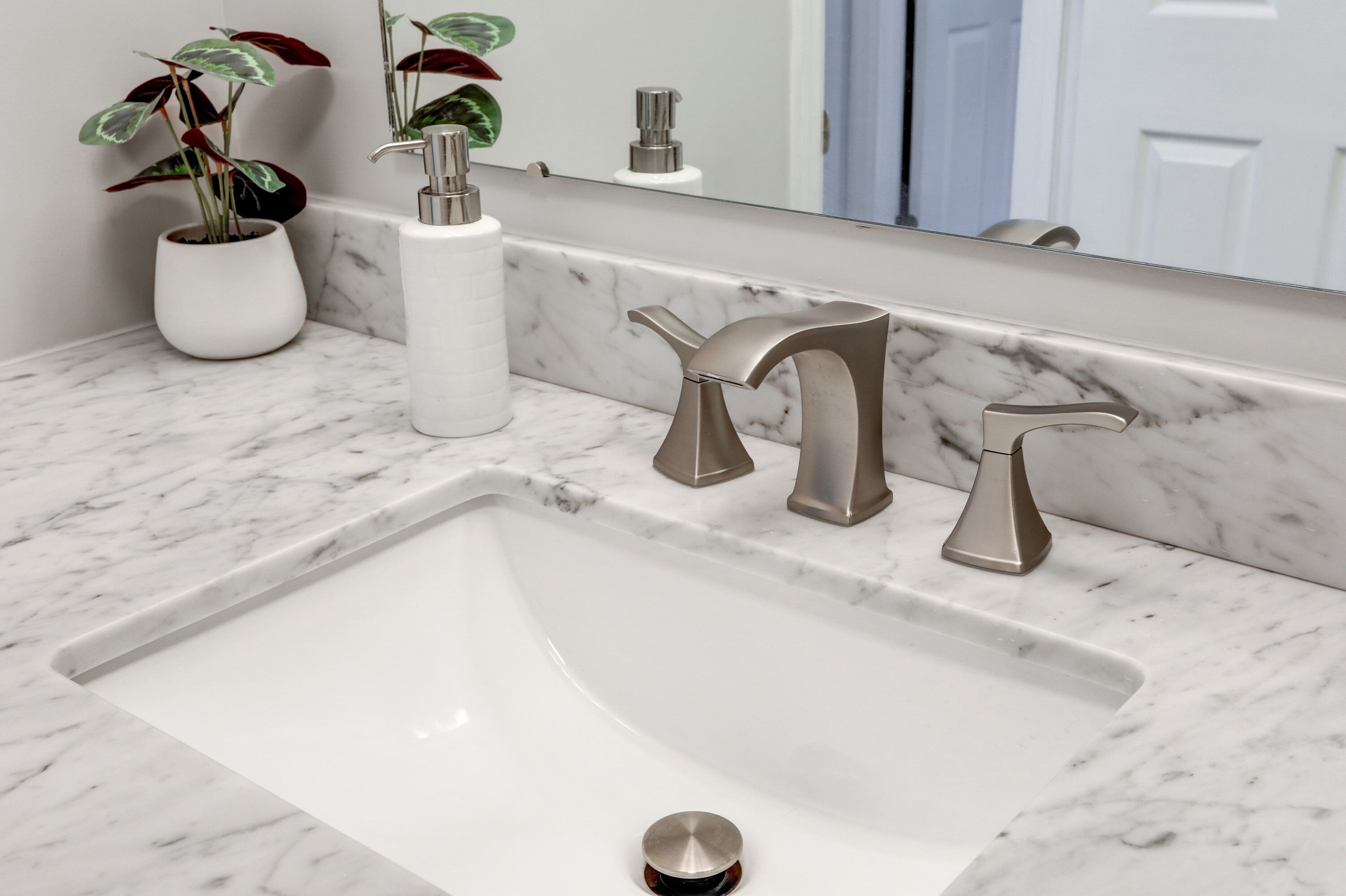 A bathroom with a white sink, two modern faucets, a marble countertop, and a potted plant.
