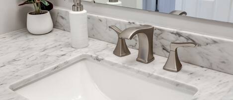 A bathroom with a white sink, two modern faucets, a marble countertop, and a potted plant.