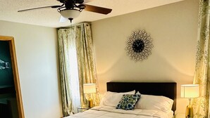 A neatly made bed with a white comforter and blue throw, two bedside lamps, a ceiling fan, and a decorative wall clock. - 5 Bedroom Private Villa with Peloton Bike (Shakopee)