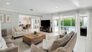 A spacious living room with a large TV, comfortable seating, and a view of a swimming pool. - 4 Bedroom Pool Home - Walk to the Beach! (West Palm Beach)