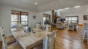 Un salón amplio con una gran mesa de comedor de madera, sillas blancas tapizadas, una chimenea de piedra y un televisor.