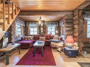 Interior de una cabaña de troncos con escalera de madera, un conjunto de sofás, una mesa de centro y una chimenea.