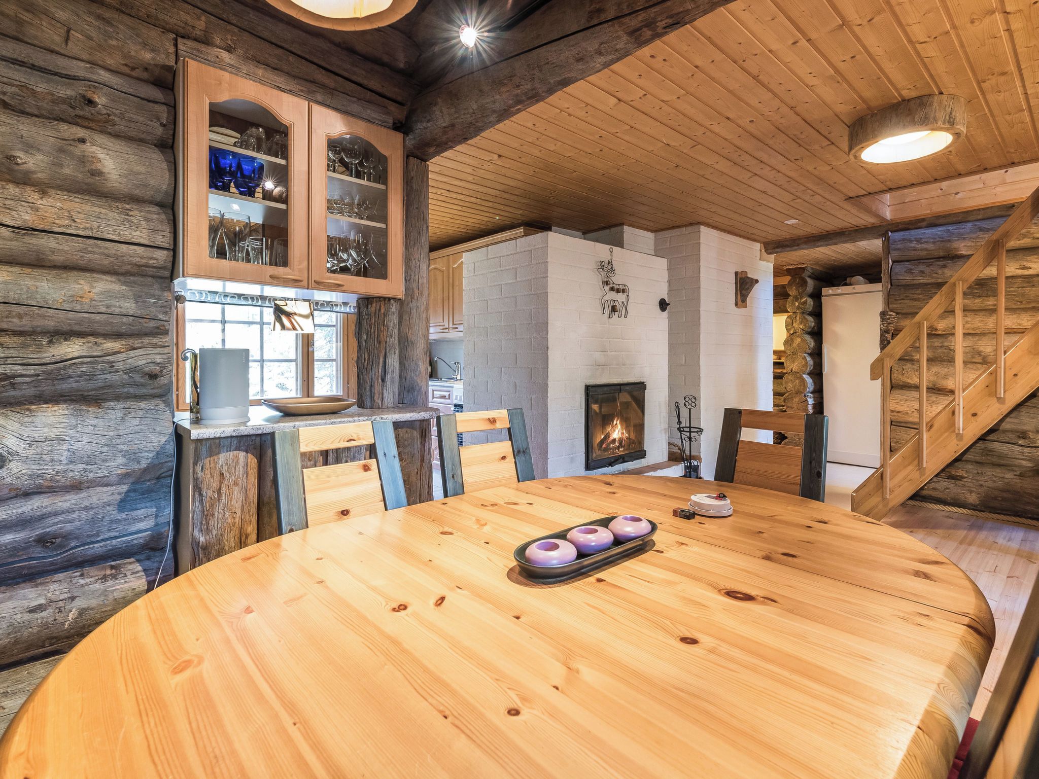 A wooden interior with a round dining table, chairs, a fireplace, and a staircase.