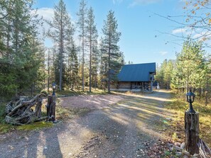 Uma cabana de toras com telhado inclinado, rodeada por uma área arborizada e um caminho de cascalho que leva à entrada.