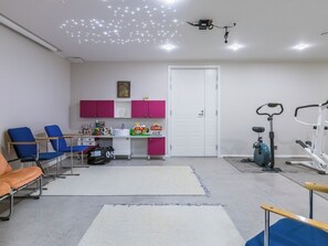 Een kamer met een bureau, stoelen, een deur, fitnessapparatuur en een plafondlamp.