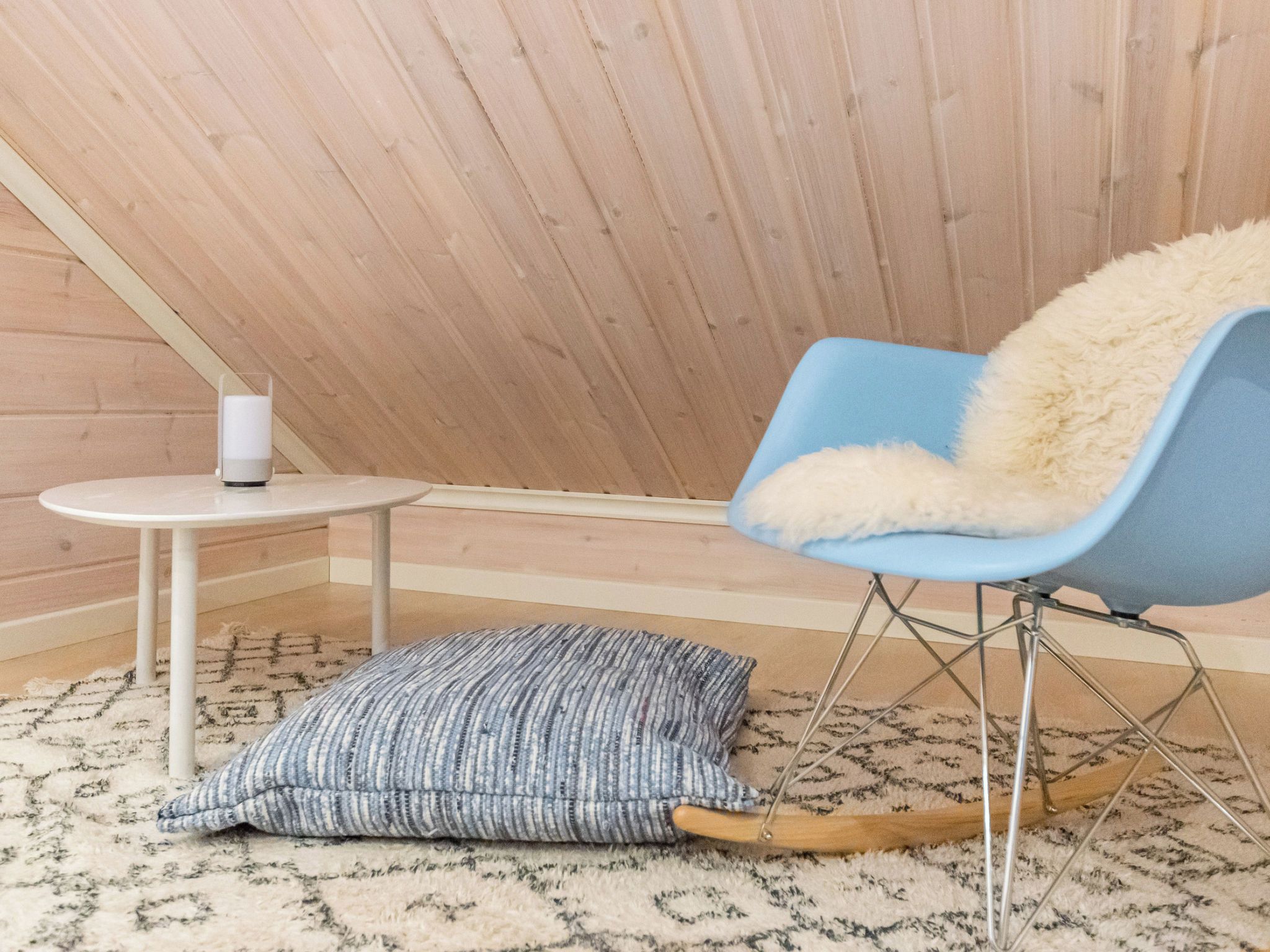 A cozy room with a sloped ceiling, a blue chair with a fluffy cushion, a small white table with a white object on it, and a patterned rug on the floor.