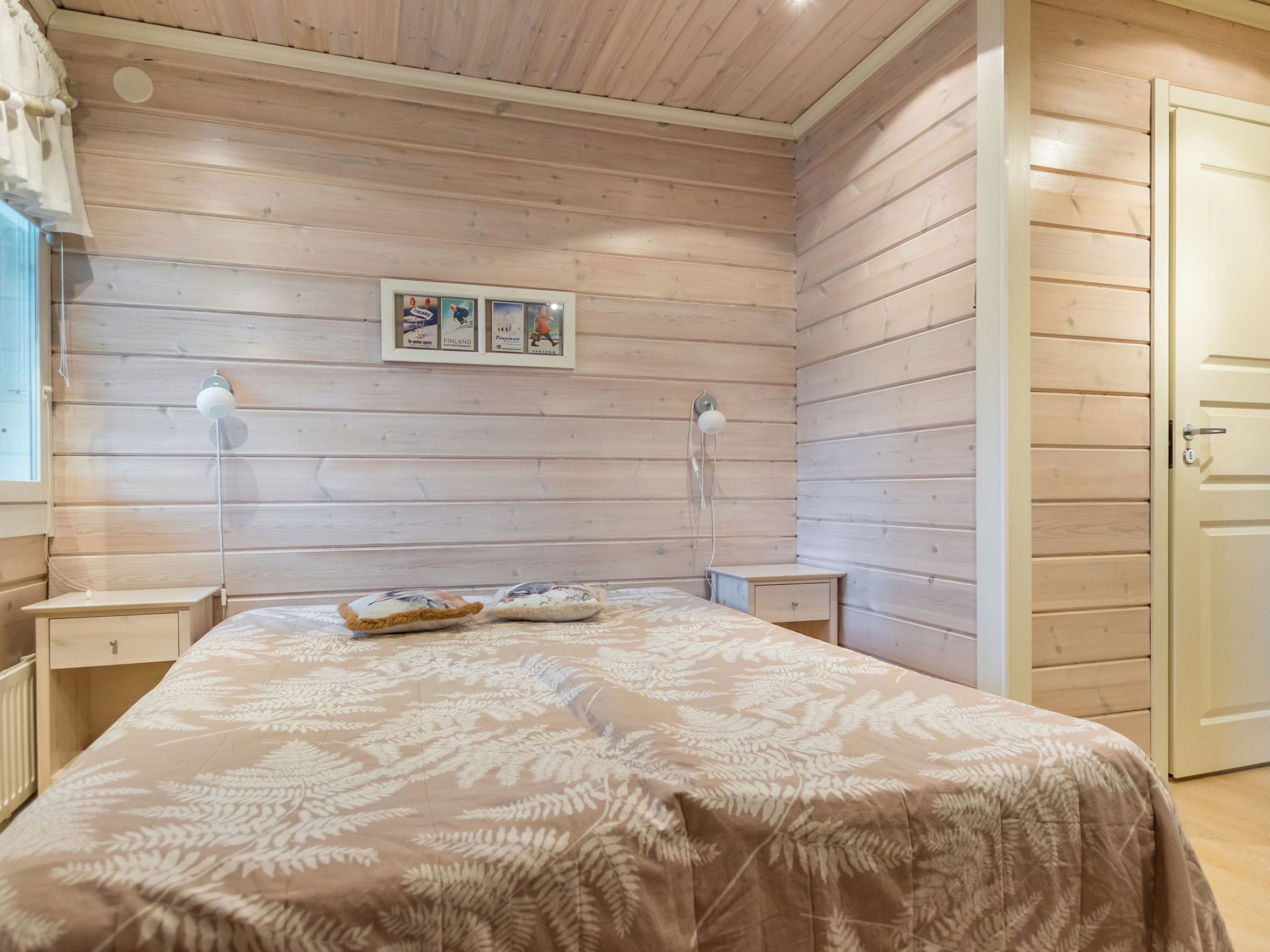 Un dormitorio con techo de listones de madera, una cama con colcha estampada con motivos de hojas, dos lámparas de noche y una puerta a la derecha.