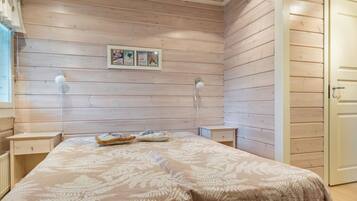 Un dormitorio con techo de listones de madera, una cama con colcha estampada con motivos de hojas, dos lámparas de noche y una puerta a la derecha.