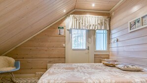 Un acogedor dormitorio en el ático con techo inclinado, paredes de madera, una cama con colcha estampada con motivos de helechos, una pequeña silla y una ventana con persianas.