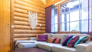 Un intérieur de chalet chaleureux avec un mur en rondins de bois, un canapé gris agrémenté de coussins colorés, une table ronde et une décoration murale en macramé.