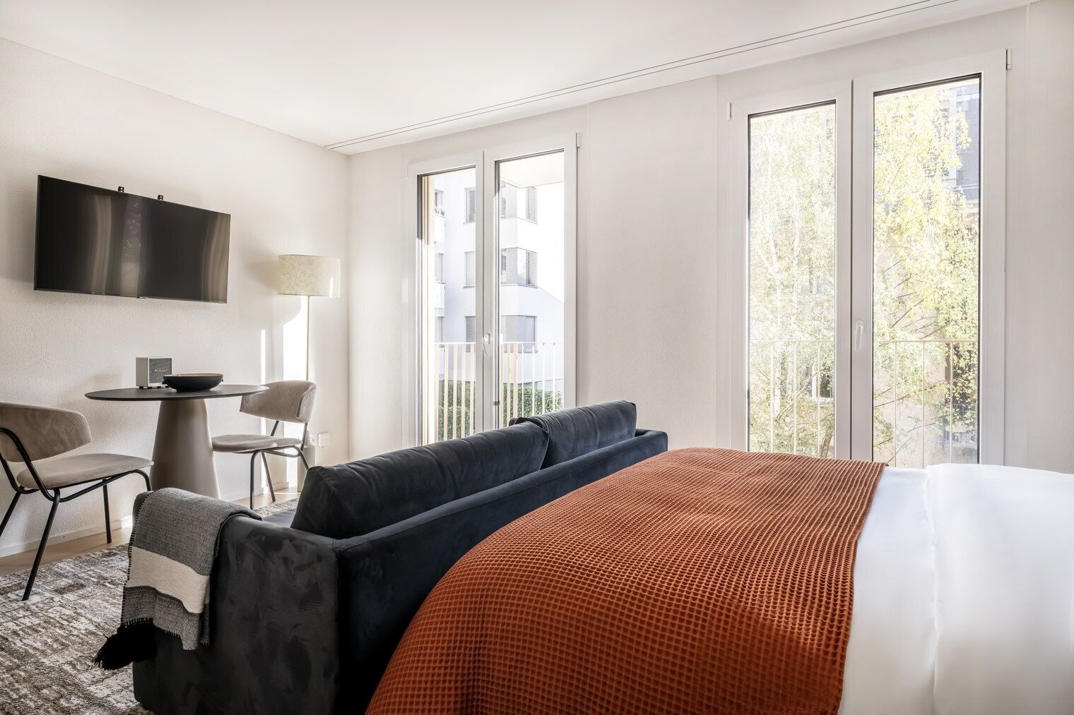Habitación de hotel con cama, sofá, escritorio, silla, televisor y ventanas amplias con vistas a edificios y árboles.