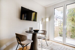 Ett modernt vardagsrum med en platt-TV som är monterad på väggen, ett runt bord med två stolar, en liten högtalare och utsikt över en stadsbild genom en stor skjutdörr i glas.
